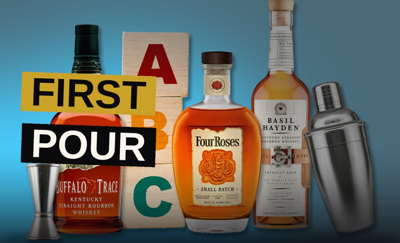 A glass of bourbon with a large ice cube sits on a wooden bar counter, with a bourbon bottle softly blurred in the background.