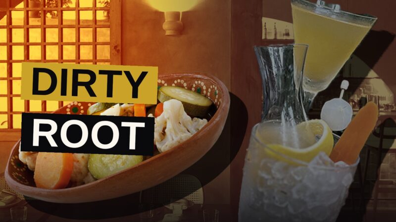 A vibrant orange Dirty Root cocktail in a glass, garnished with a lemon twist, pickled carrot, and cocktail onion.