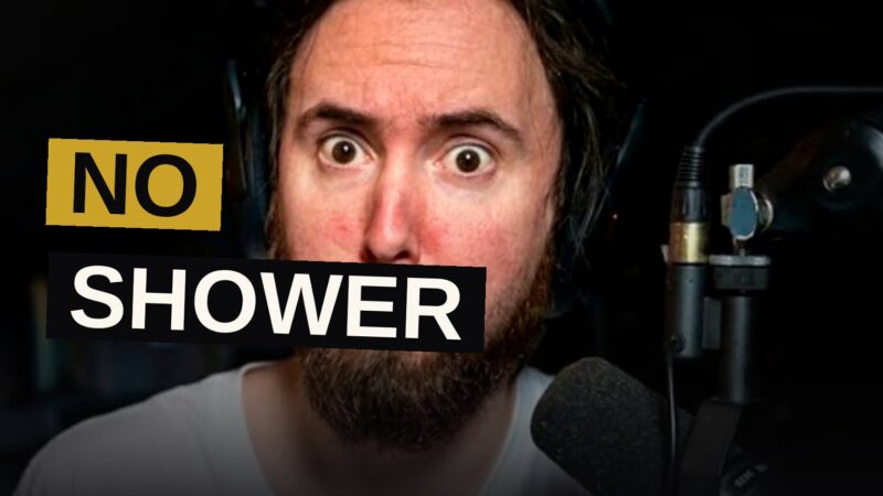 Asmongold sits at his gaming desk with messy hair, looking unbothered, in a dimly lit room with RGB lighting.