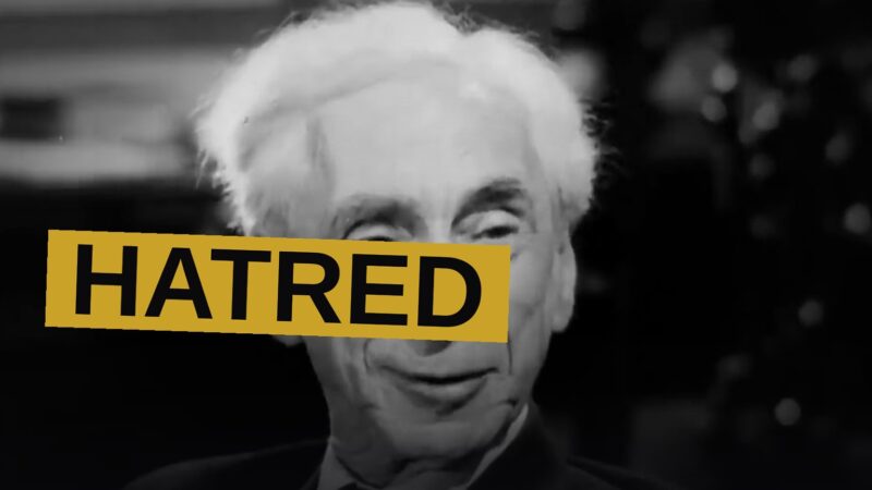 An aged philosopher, seated before old cameras, speaks with grave conviction upon a shadowed stage.