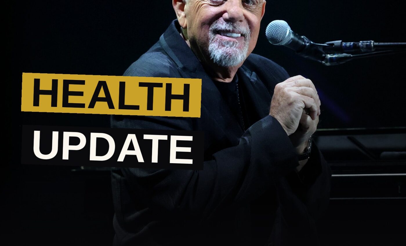 Billy Joel smiles gently in a dimly lit studio, wearing sunglasses and a casual shirt.