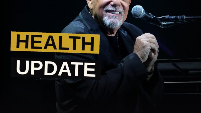 Billy Joel smiles gently in a dimly lit studio, wearing sunglasses and a casual shirt.