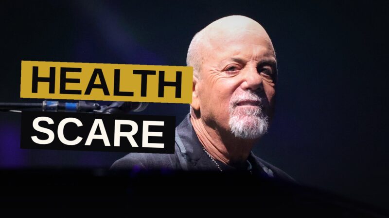 Billy Joel sits alone backstage in a dimly lit room, looking thoughtful and concerned.