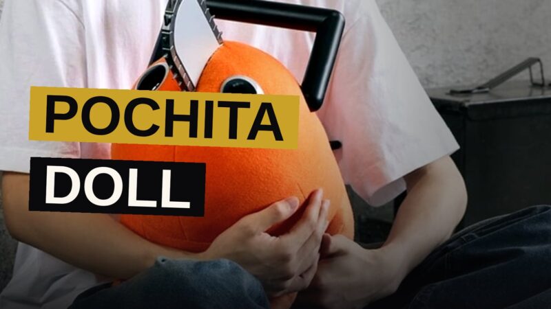 A life-size orange Pochita doll with a chainsaw blade sits posed on a wooden floor in a bright room.
