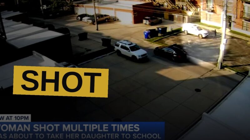 A black sedan drives away from a residential building with a shattered balcony window and a bullet-riddled car parked outside, police tape in the foreground.