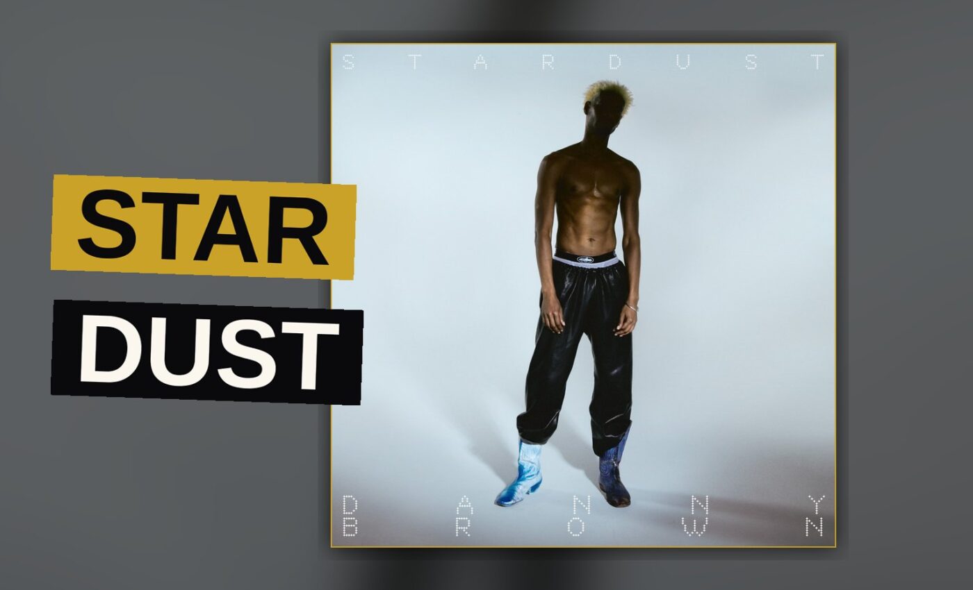 A rapper stands under pink and gold stage lights as dust swirls around him.
