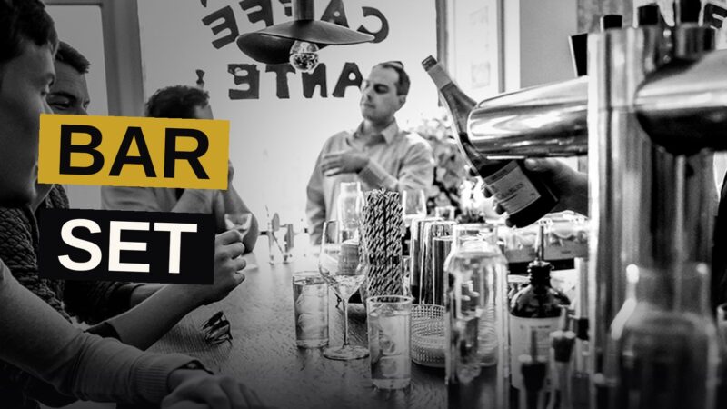 A bartender in a Dante apron slides two drinks across a bar counter inside a stylish, warmly lit New York City bar.
