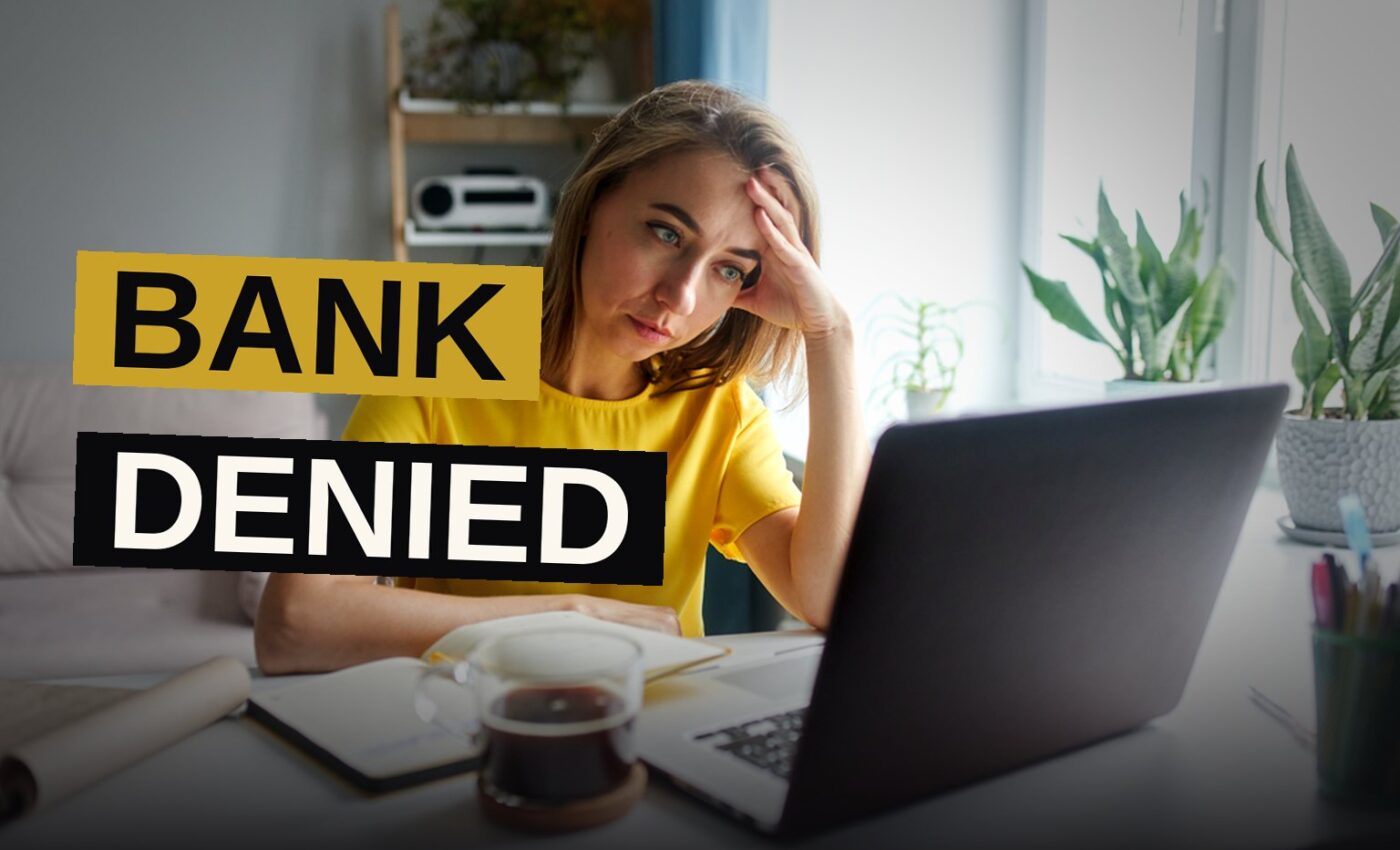 A person holding a bank account denial letter while sitting at a desk with a laptop and paperwork.