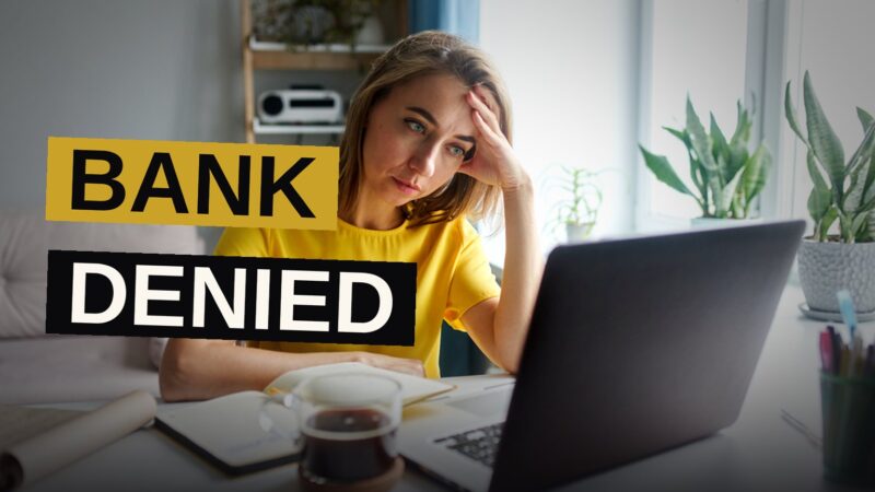 A person holding a bank account denial letter while sitting at a desk with a laptop and paperwork.