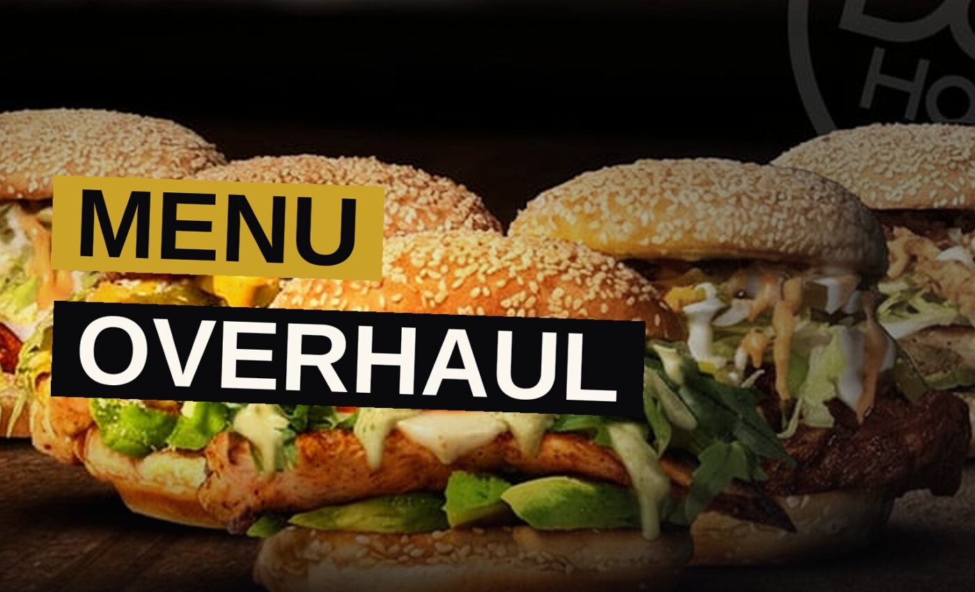 An overhead view of a gourmet hot dog, grilled chicken sandwich, and loaded tot-chos on a wooden table.