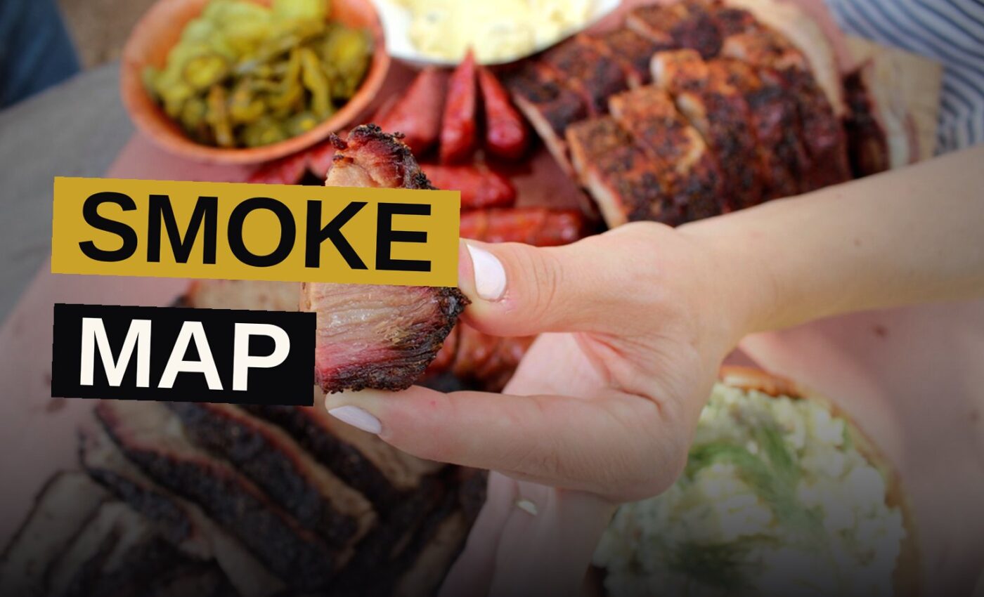 A wooden board piled with sliced brisket, ribs, and sausages, served with barbecue sauce and pickles.