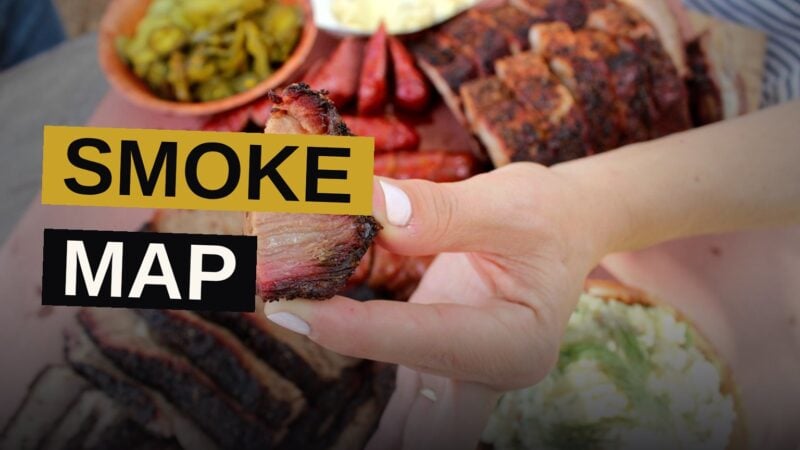 A wooden board piled with sliced brisket, ribs, and sausages, served with barbecue sauce and pickles.