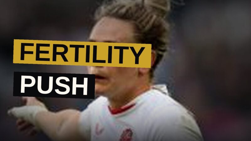 A rugby player stands confidently on a sunlit field, her expression showing determination.