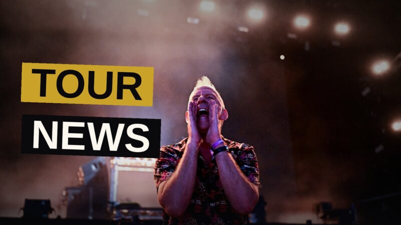 A DJ performs on stage under vivid lights at a packed warehouse, with the crowd's hands raised in the air.