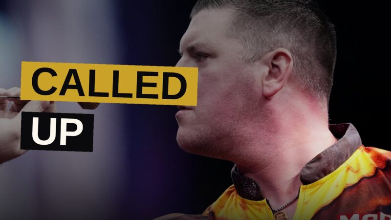 A professional darts player throws a dart during a tournament under stage lighting.
