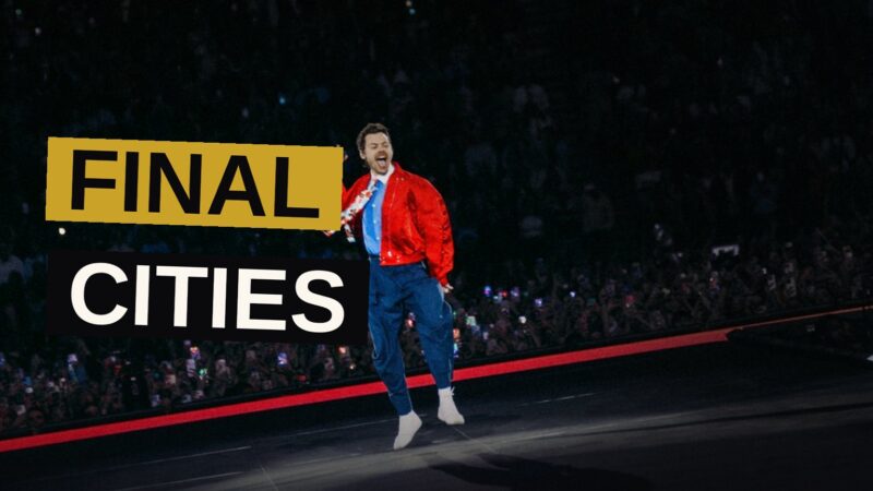 Harry Styles performs on stage during a concert, with fans' hands raised and bright lights illuminating the arena.