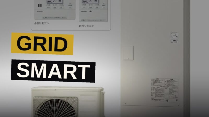 A modern white heat pump water heater unit outside a house with solar panels on the roof.