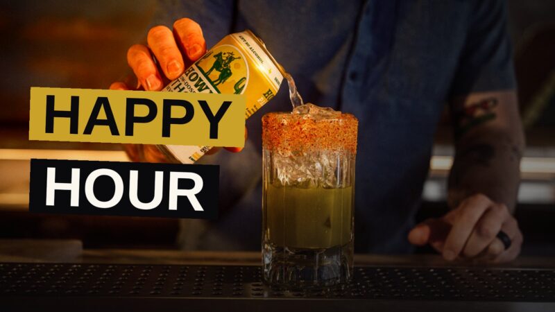 A flat-lay of colorful cocktails and small plates on a wooden table with a blurred neon cityscape in the background.