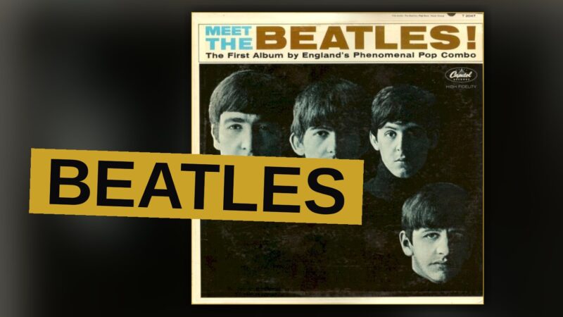 The Beatles posing in matching dark suits with mop-top haircuts, looking at the camera with confident expressions.