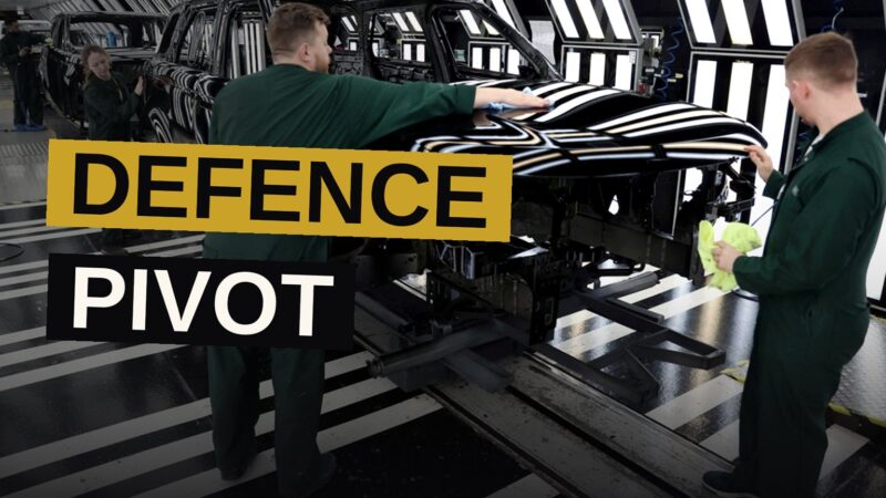 Workers in an automotive factory pause near a production line, with a faint military vehicle silhouette visible in the background.