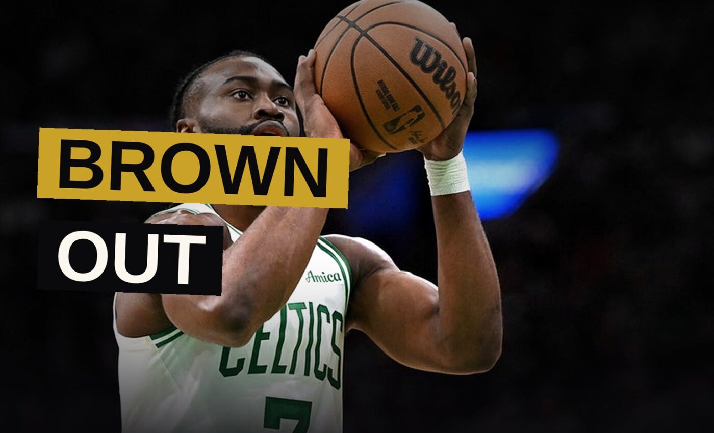 Jaylen Brown stands in a shadowed arena, arms crossed, wearing a basketball jersey, with a thoughtful expression.