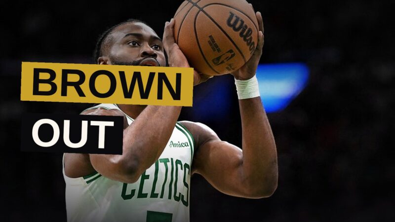 Jaylen Brown stands in a shadowed arena, arms crossed, wearing a basketball jersey, with a thoughtful expression.