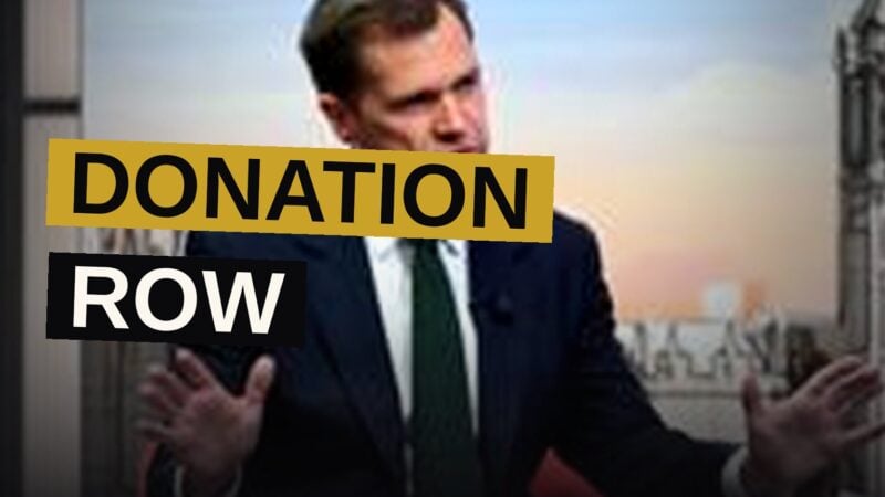 A politician speaks at a press conference with a large screen displaying the figure '£72m' behind him.