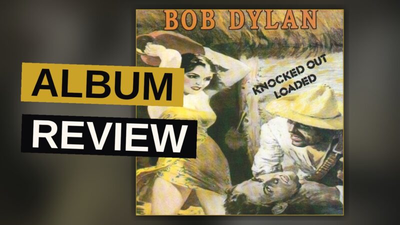 A vintage cassette tape of Bob Dylan's album Knocked Out Loaded rests on a wooden table in a dimly lit room.