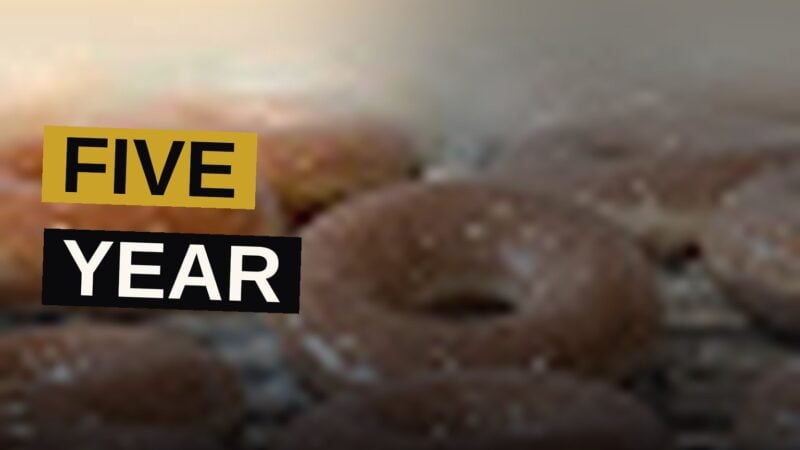 An apple cider glazed donut sits among a dozen football-shaped donuts on a bakery plate.