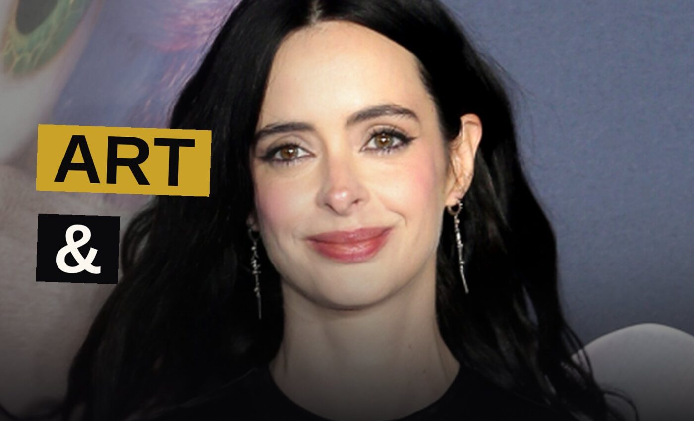 Krysten Ritter stands in a dark studio, holding a paintbrush, with colorful paint strokes around her.