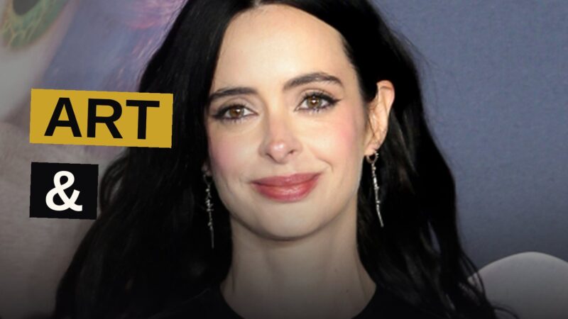 Krysten Ritter stands in a dark studio, holding a paintbrush, with colorful paint strokes around her.