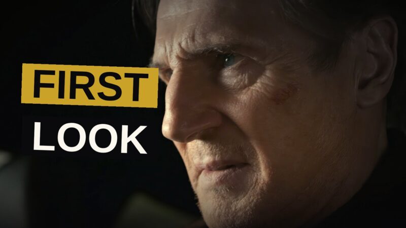 Liam Neeson stands confidently in front of a sleek car as police cars and a helicopter pursue him in the background.