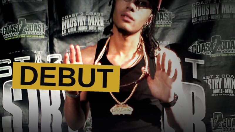 A young rapper stands in a dim room surrounded by vinyl records, evoking the era of his debut album.