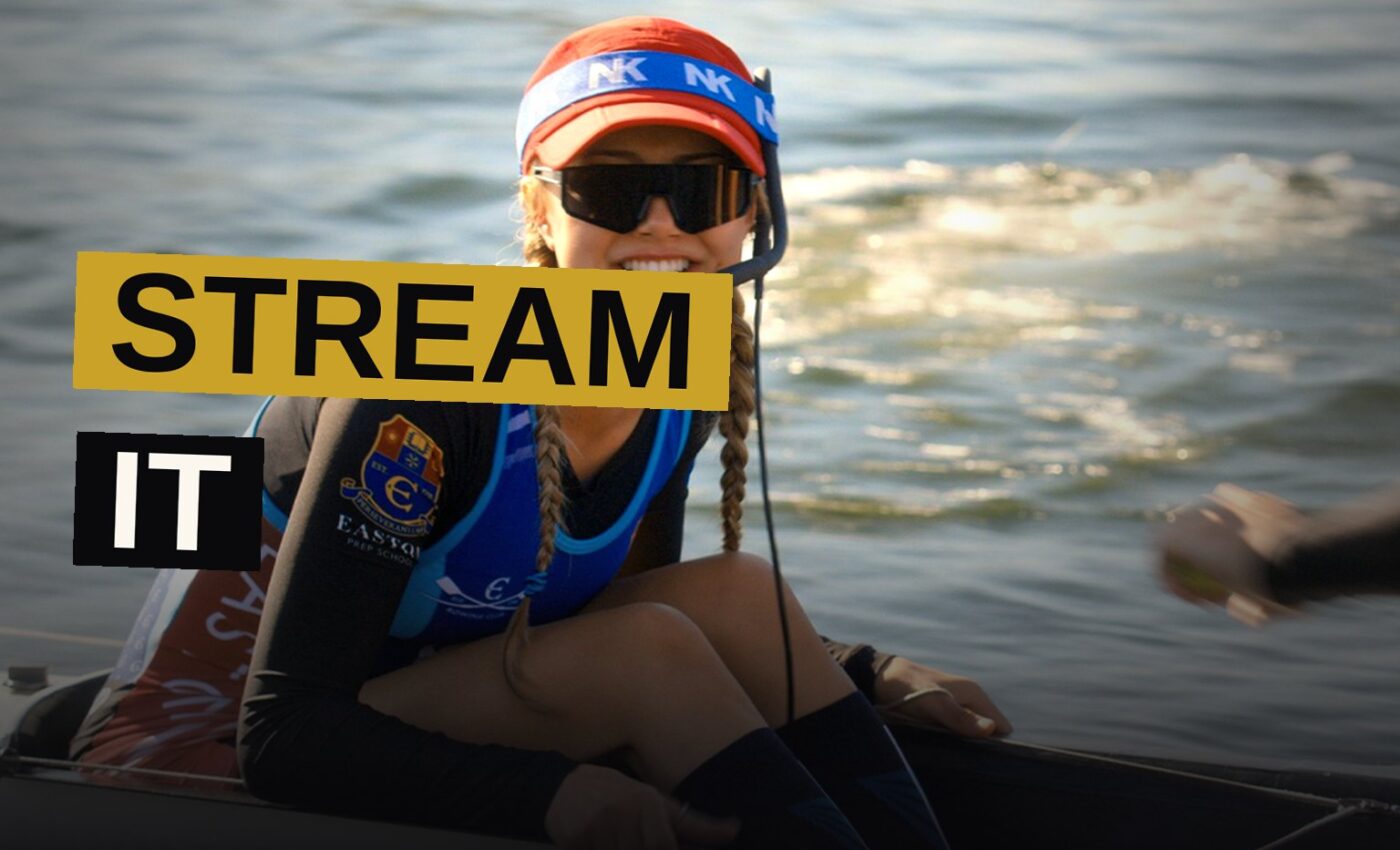 A teenage girl coxswain stands confidently at the front of a rowing boat with her male team on a river at sunset.