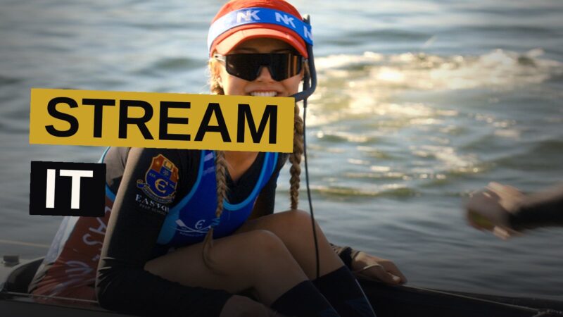 A teenage girl coxswain stands confidently at the front of a rowing boat with her male team on a river at sunset.