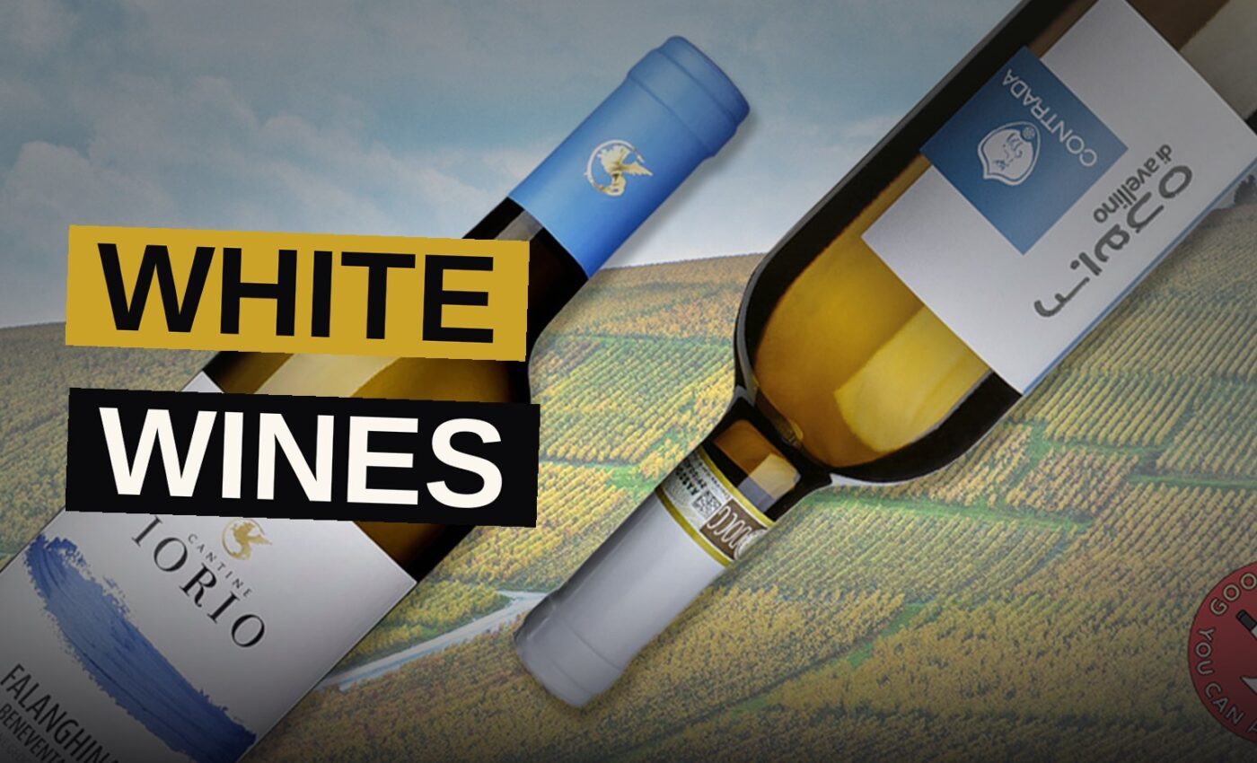 A collection of white wine bottles and glasses arranged on a table with an Italian vineyard landscape in the background.