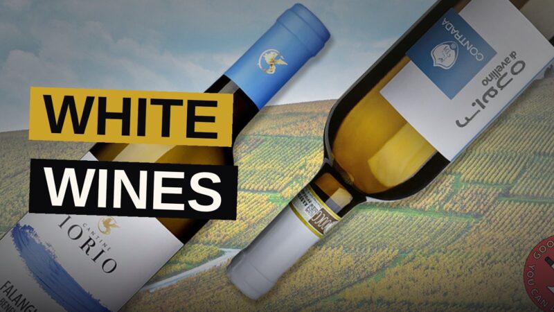 A collection of white wine bottles and glasses arranged on a table with an Italian vineyard landscape in the background.