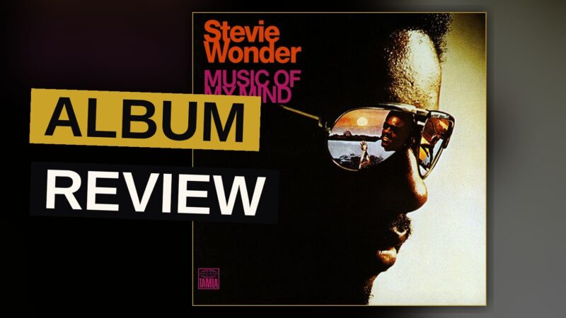 A young Stevie Wonder in the 1970s, smiling, surrounded by keyboards and synthesizers in a recording studio.