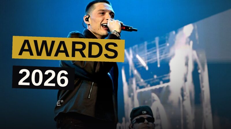 A singer performs on a brightly lit award show stage with golden light beams.