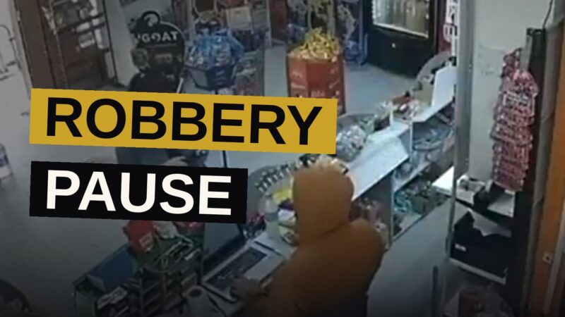A hooded man in a store hands a receipt to a customer while another hooded figure stands by the entrance, in a CCTV-style image.