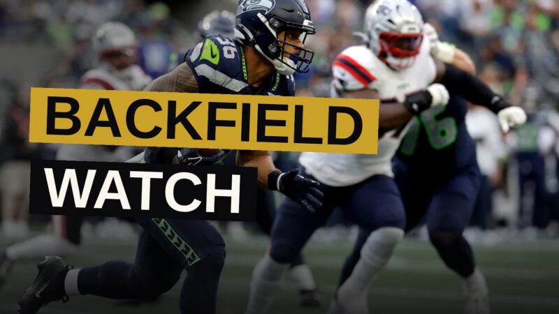 A Seattle Seahawks running back carrying the football during a game, with the stadium blurred in the background.