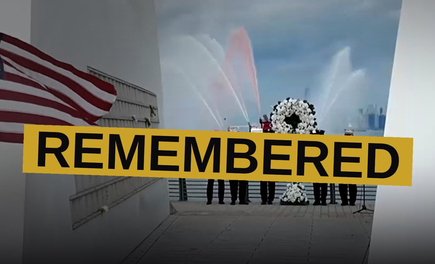 A memorial fence with American flags and firefighters' names outside a stadium, with a fireboat spraying water in the background.
