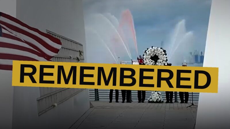 A memorial fence with American flags and firefighters' names outside a stadium, with a fireboat spraying water in the background.