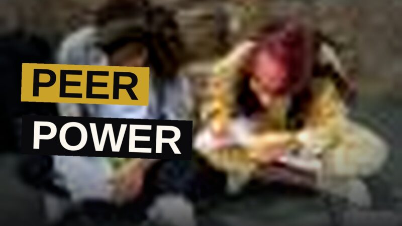 A teenage girl teaches younger students coding at a table with laptops in a bright classroom.