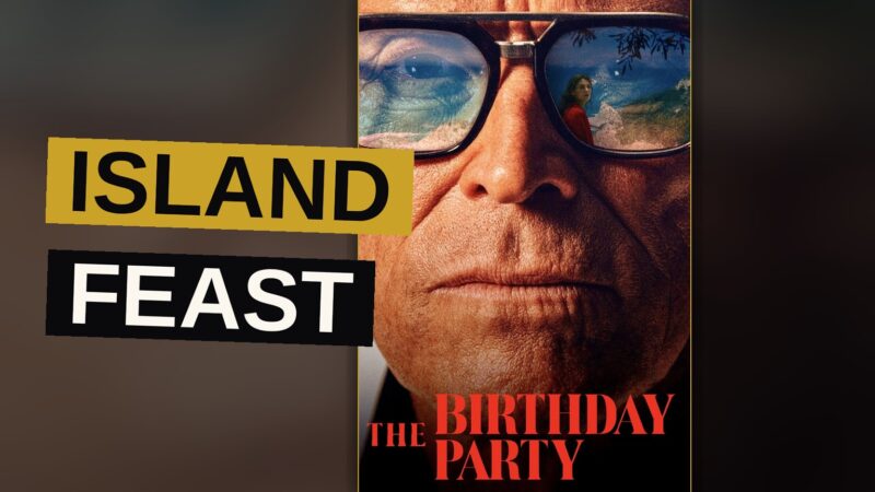 A wealthy man stands apart from his guests at a lantern-lit island feast, his face heavy with unspoken weariness.