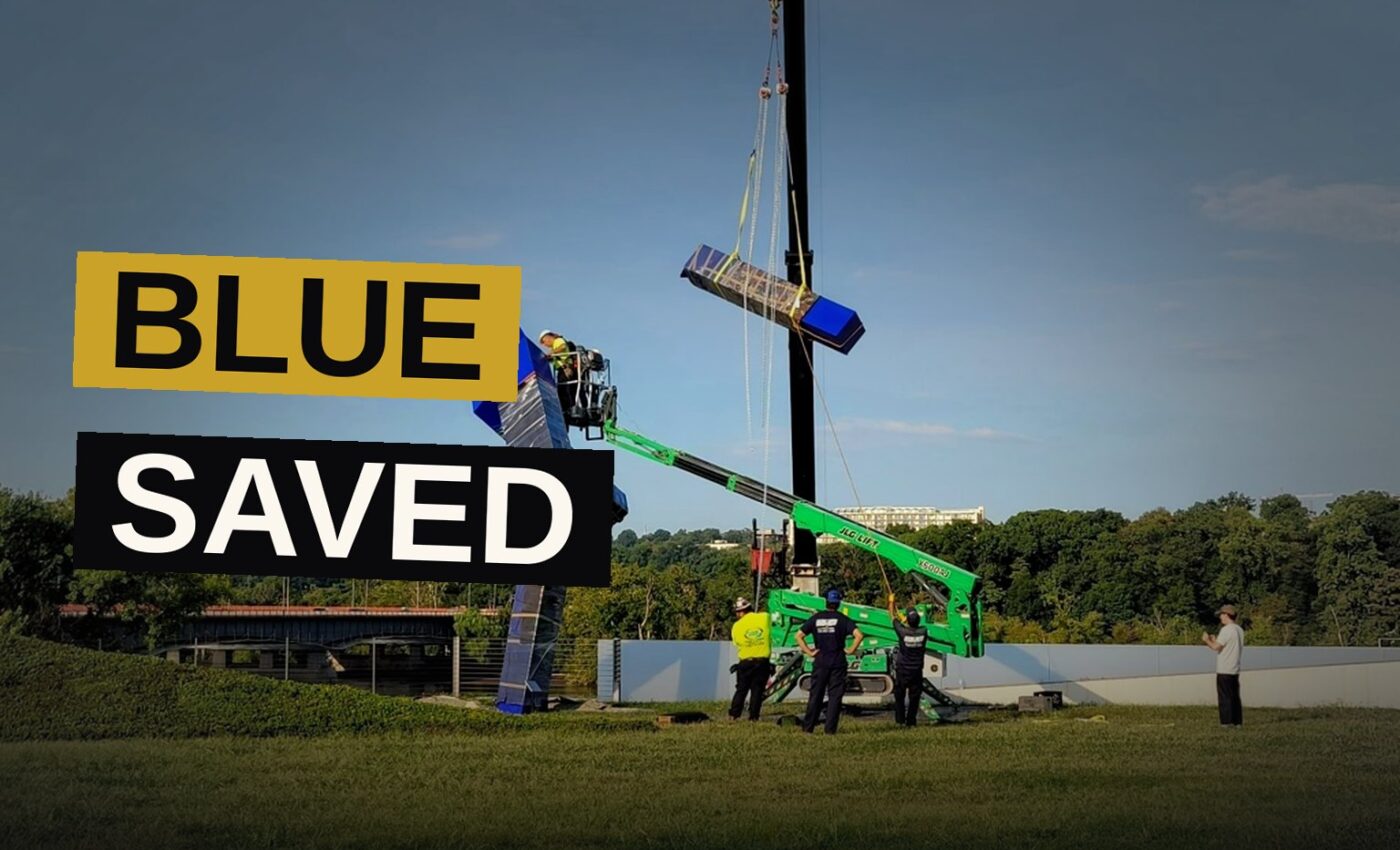 A tall blue sculpture, once a gift of honor, is lifted away from its former resting place beneath a heavy sky.