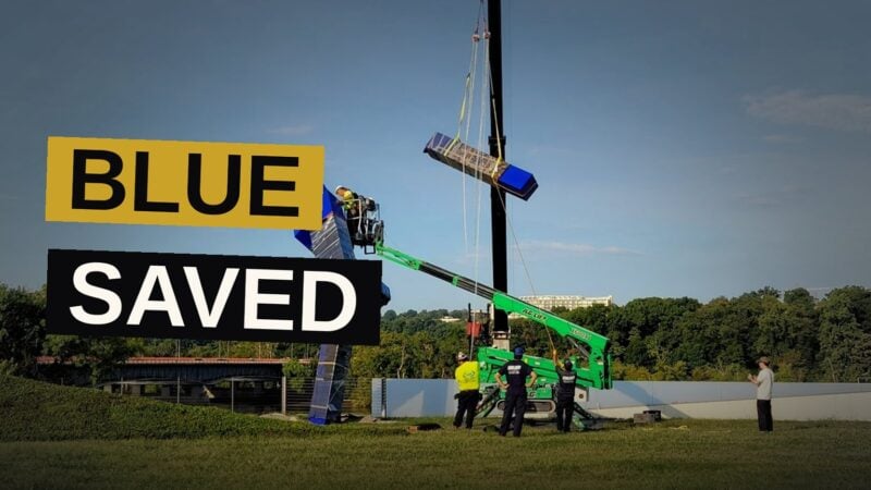 A tall blue sculpture, once a gift of honor, is lifted away from its former resting place beneath a heavy sky.