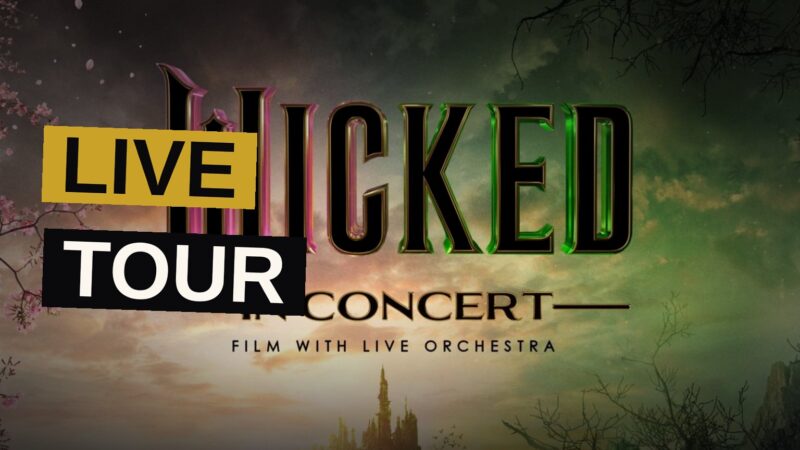A symphony orchestra performs live in a concert hall beneath a giant screen showing a scene from Wicked.
