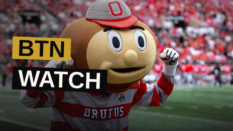 A smartphone displays a live broadcast of a college football game beneath a glowing stadium scoreboard.